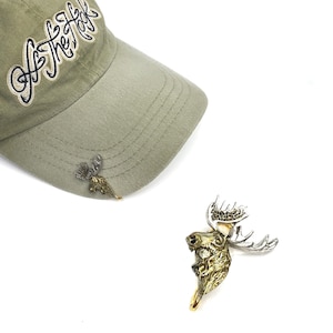 May include: A green baseball cap with the text "Of The Hook" embroidered on the front. The cap has a moose pin attached to the brim. A second moose pin is shown separately.