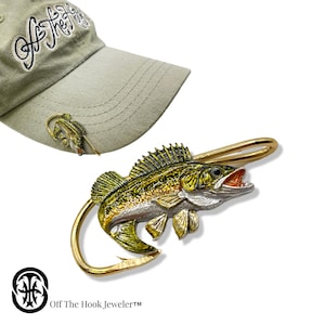 May include: A gold-colored fishing lure hat clip featuring a detailed walleye fish with green, yellow, and white markings. The clip is attached to a baseball cap with the text "Off The Hook, Jeweler™" on the front.