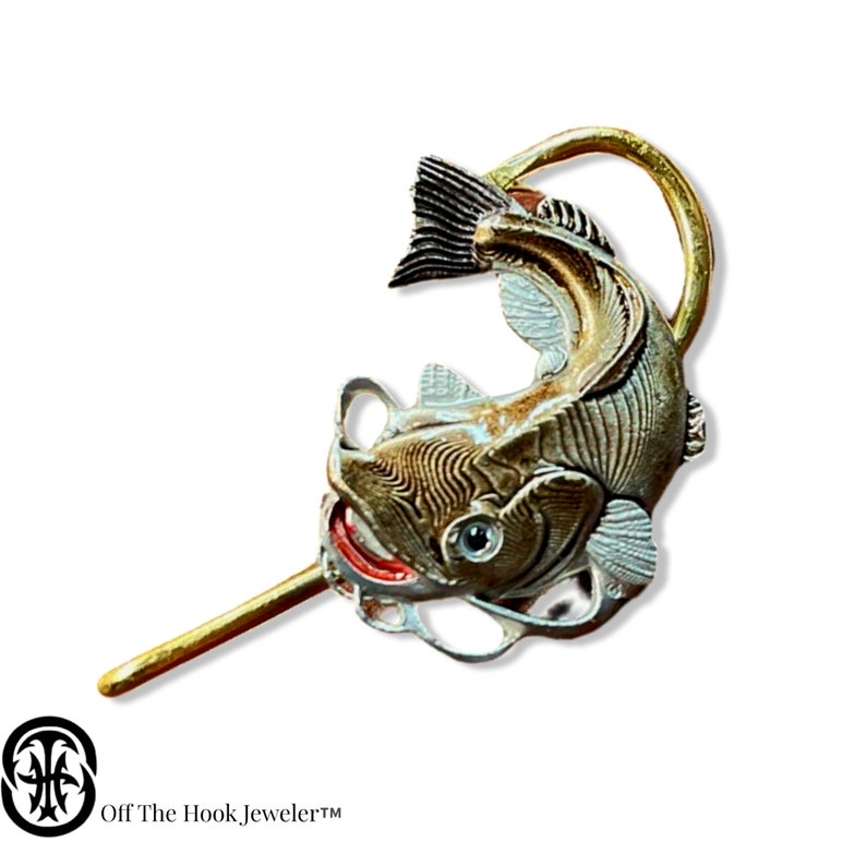 May include: A gold and silver fish pin with a detailed design. The fish is depicted with its mouth open and its tail curled up. The pin is attached to a thin gold rod.