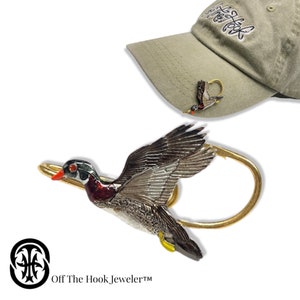 May include: A gold-tone metal pin featuring a detailed, hand-painted wood duck in flight. The pin is designed to attach to a hat or other fabric. The pin is shown attached to a green baseball cap with the text "Off The Hook" embroidered on the front.