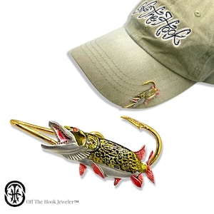 May include: A gold-tone fishing lure pin with a detailed, realistic design of a pike fish. The pin is attached to a hat with the text "Off The Hook, Jeweler" on the hat.
