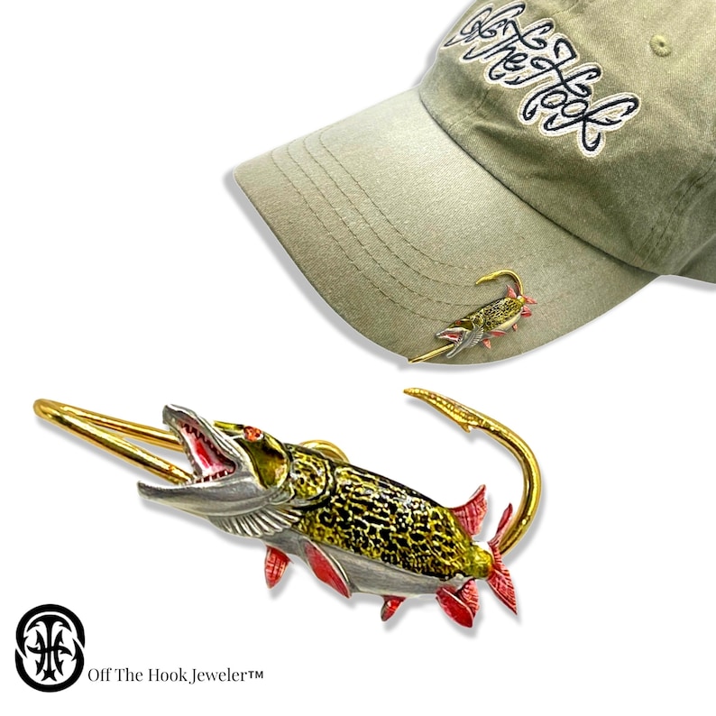 May include: A gold-tone fishing lure pin with a detailed, realistic design of a pike fish. The pin is attached to a hat with the text "Off The Hook, Jeweler" on the hat.