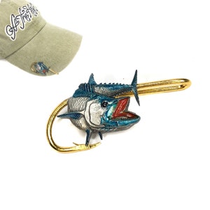 May include: A gold-colored fish hook with a blue and silver fish attached. The fish has an open mouth with red inside. The hook is attached to a gold-colored bar.