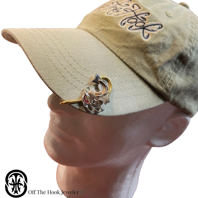 May include: A silver and gold fishing lure shaped like a bass with a red mouth, attached to a gold hat clip. The clip is attached to a green baseball cap with the words "Off The Hook" embroidered on the front.