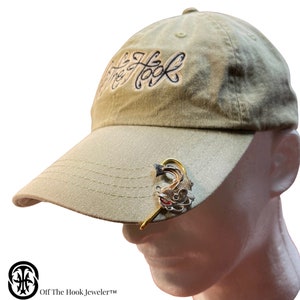 May include: A beige baseball cap with the text "Off The Hook" embroidered on the front. A gold-colored fish hook with a fish charm is attached to the brim of the cap.