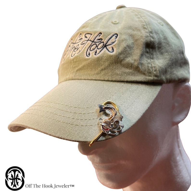 May include: A beige baseball cap with the text "Off The Hook" embroidered on the front. A gold-colored fish hook with a fish charm is attached to the brim of the cap.