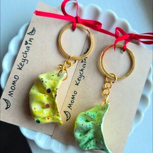 May include: Two handmade keychains featuring dumpling charms in yellow and green. Each keychain has a gold-colored ring and a small card with the text "Momo Keychain" and a red ribbon.