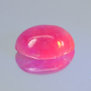 May include: A polished, oval-shaped gemstone in a vibrant shade of pink. The gem is smooth and reflective, with a glossy surface. The image shows the gem's reflection, creating a mirrored effect. The gemstone is likely used in jewelry.