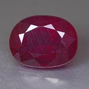 Può includere: Una pietra preziosa di colore rosso intenso e forma ovale con una superficie sfaccettata. La pietra mostra una qualità traslucida con inclusioni interne visibili e una finitura lucida, che riflette la luce attraverso le sue molteplici sfaccettature.