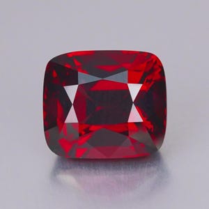 Puede incluir: Una piedra preciosa de color rojo intenso, con talla cojín y una superficie facetada que crea un patrón geométrico de reflejos claros y oscuros. La piedra tiene forma cuadrada con esquinas redondeadas y un color carmesí vibrante y transparente.