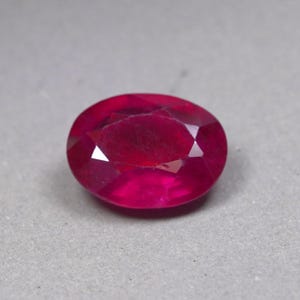 May include: A loose, oval-cut gemstone with a deep, vibrant red color. The stone features multiple triangular and trapezoidal facets across its surface, creating a reflective, polished appearance that catches light from various angles.