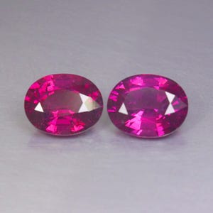 Pode incluir: Duas pedras preciosas de corte oval com uma cor magenta-rosa profunda e vibrante. Cada pedra apresenta uma superfície facetada que reflete a luz, criando uma aparência brilhante e cintilante. As gemas estão posicionadas lado a lado.