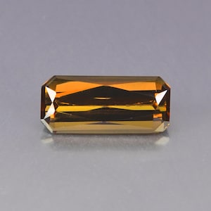 May include: A rectangular, faceted gemstone with a warm, amber-orange hue. The gem's cut features a series of parallel facets, creating a play of light and shadow. The gemstone is set against a neutral gray background.