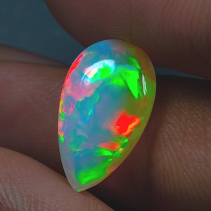 Puede incluir: Una piedra preciosa de ópalo en forma de pera con colores vibrantes. El ópalo muestra un juego de colores con destellos de tonos rojos, verdes y azules. La piedra se sostiene entre los dedos, mostrando su superficie pulida y belleza natural.