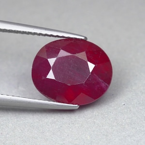 Può includere: Una gemma sfaccettata di forma ovale color rosso intenso, tenuta da una pinzetta metallica color argento. La pietra presenta un taglio simmetrico con molteplici superfici piatte che riflettono la luce, mostrando un colore cremisi ricco e saturo.