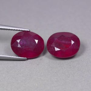 May include: Two oval-cut, deep red gemstones with faceted surfaces. One gemstone is held by silver-toned metal tweezers, while the other sits beside it. The stones exhibit a translucent quality with visible internal inclusions and a polished finish.
