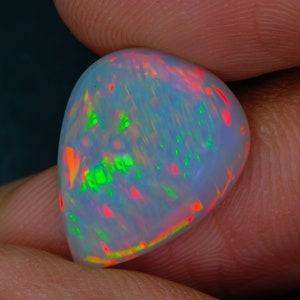 May include: A pear-shaped cabochon opal gemstone featuring a translucent white base with vibrant flashes of neon green and bright red play-of-color. The stone has a smooth, polished surface and a rounded, teardrop-like silhouette.