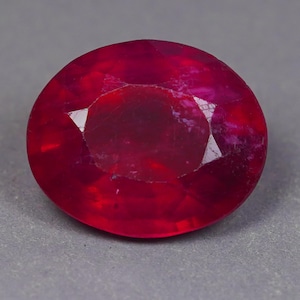 Puede incluir: Una gema de rubí de corte ovalado vibrante con un color rojo profundo y saturado. La piedra muestra una superficie facetada con múltiples planos geométricos que reflejan la luz, creando una apariencia brillante en toda su estructura pulida y translúcida.
