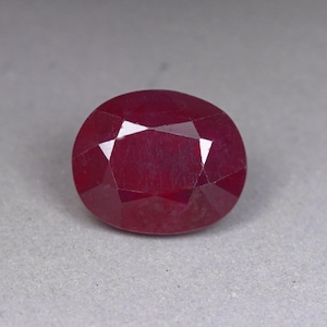May include: A deep red, oval-shaped gemstone with a faceted surface. The stone displays a rich, saturated crimson hue and a polished finish, with multiple triangular and trapezoidal facets reflecting light across its translucent, rounded body.