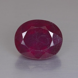 Puede incluir: Una piedra preciosa de color rojo intenso con forma ovalada y superficie facetada. La piedra muestra una calidad translúcida con inclusiones internas visibles y un acabado pulido, reflejando la luz a través de sus múltiples facetas geométricas.