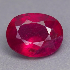 Könnte beinhalten: Ein leuchtender, oval geschliffener Rubin-Edelstein mit einer tiefen, gesättigten roten Farbe und facettierten Oberflächen, die das Licht reflektieren. Der Stein weist interne Einschlüsse und eine polierte Oberfläche auf und zeigt eine klassische, symmetrische ovale Form.