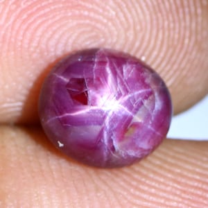 Może przedstawiać: Wypolerowany, okrągły kamień szlachetny o szlifie kaboszonowym z efektem gwiazdy. Klejnot ma głęboki czerwonofioletowy kolor, z sześcioramiennym wzorem gwiazdy widocznym na jego powierzchni. Klejnot jest trzymany między palcami.