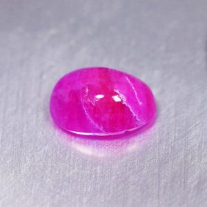 May include: A vibrant, oval-shaped gemstone with a polished, smooth surface. The stone displays a deep, saturated magenta-pink color with subtle internal white veining and light reflections across its rounded, cabochon-style top surface.