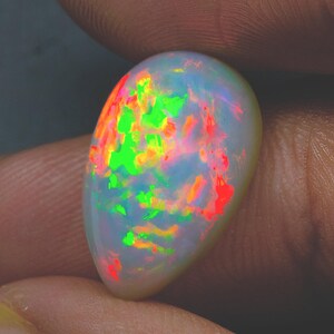 May include: A pear-shaped cabochon opal gemstone featuring a translucent white base with vibrant flashes of neon green, bright orange, and red play-of-color throughout the surface, displaying a smooth, polished, and rounded dome-like structure.