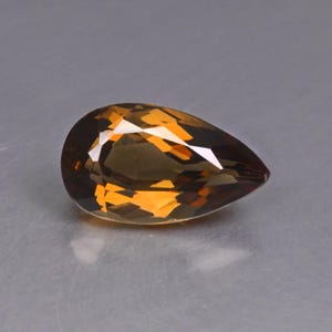 Könnte beinhalten: Ein birnenförmiger, facettierter Edelstein in tiefem Goldbraun. Der Stein besitzt einen Brillantschliff mit zahlreichen dreieckigen und rautenförmigen Facetten, die das Licht reflektieren und ein kontrastreiches Muster aus dunklen und hellen Bernsteintönen erzeugen.