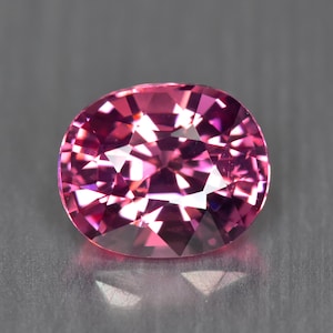 Puede incluir: Una gema facetada, de forma ovalada, con un tono rosa vibrante. La superficie de la gema refleja la luz, creando un efecto brillante. La gema se presenta sobre un fondo gris, resaltando su color y brillo. Ideal para joyería.