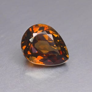 Peut inclure: Une pierre précieuse en forme de poire présentant une palette de couleurs ambre profond et brun doré. La pierre affiche une surface facettée avec des coupes géométriques complexes qui reflètent la lumière, créant un aspect contrasté.