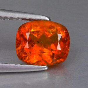 Könnte beinhalten: Ein feueropal-Edelstein im Kissenschliff mit leuchtend orangefarbenem Ton und inneren gelben und roten Lichtreflexen. Der Edelstein wird von einer silberfarbenen Metallpinzette gehalten und zeigt seine facettierte Oberfläche und Leuchtkraft.
