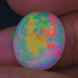 May include: An oval-shaped opal gemstone with a vibrant display of colors. The opal features a mix of green, yellow, orange, and pink hues, creating a mesmerizing play of light. The gemstone is held between fingers.