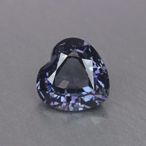 May include: A heart-shaped, faceted gemstone with a deep blue-violet hue. The gem has a polished surface that reflects light, creating a sparkling effect. The stone is set against a neutral gray background.