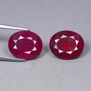 May include: Two oval-cut, deep red rubies with faceted surfaces. One ruby is held by silver-toned metal tweezers, while the other sits independently. Both gemstones exhibit a vibrant, saturated crimson hue and a polished, reflective finish.