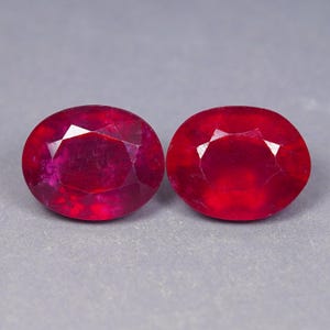 Pode incluir: Dois rubis de corte oval com uma cor vermelha profunda e superfícies facetadas. As pedras preciosas exibem uma qualidade translúcida com inclusões internas e um acabamento polido, refletindo a luz através das suas formas simétricas e arredondadas.