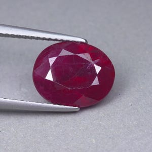 May include: An oval-cut, deep red ruby gemstone with a faceted surface, held by a pair of silver-toned metal tweezers. The stone displays a rich, saturated crimson hue with visible internal inclusions and a polished, reflective finish.
