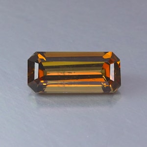 Peut inclure: Une pierre précieuse rectangulaire à taille émeraude présentant une palette de couleurs ambre profond et brun doré. La pierre présente des facettes nettes et propres qui créent un motif géométrique de reflets clairs et sombres.