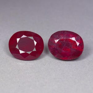 Può includere: Due rubini di forma ovale color rosso intenso con superfici sfaccettate. Il rubino a sinistra mostra un chiaro motivo di taglio geometrico, mentre il rubino a destra presenta una struttura interna più naturale e testurizzata.