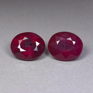 Può includere: Due rubini sfaccettati di forma ovale di colore rosso intenso. Ogni gemma presenta una superficie lucida con molteplici sfaccettature triangolari che riflettono la luce, mostrando una qualità traslucida e una ricca tonalità cremisi satura.