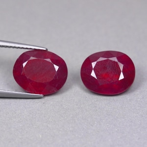 Puede incluir: Dos rubíes de color rojo intenso con corte ovalado y superficies facetadas. Un rubí está sujeto por unas pinzas de metal plateado, mientras que el otro descansa de forma independiente. Ambas piedras preciosas muestran un tono carmesí vivo.