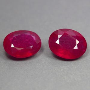 Op de afbeelding: Twee ovaal geslepen robijnen met een dieprode kleur en gefacetteerde oppervlakken. Deze edelstenen vertonen een briljante glans en heldere transparantie, met een klassieke ovale vorm en meerdere driehoekige facetten die het licht reflecteren.