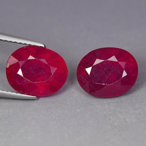 May include: Two oval-cut, deep red gemstones with faceted surfaces. One gemstone is held by silver-toned metal tweezers, while the other sits independently. Both stones exhibit a vibrant, saturated crimson hue and a polished, reflective finish.
