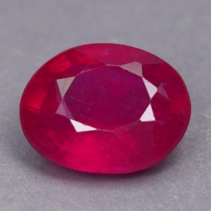 Può includere: Una gemma di rubino dal taglio ovale con una tonalità rossa profonda e satura. La pietra presenta una superficie sfaccettata con una tavola centrale e tagli geometrici simmetrici che riflettono la luce, creando un aspetto brillante.