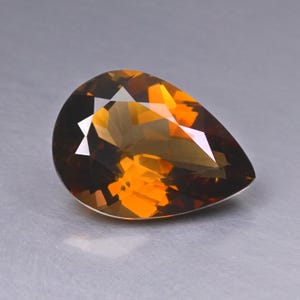 Könnte beinhalten: Ein facettierter, birnenförmiger Edelstein mit einem Farbverlauf von tiefbraun an den Rändern zu leuchtendem Orange in der Mitte. Der Schliff erzeugt mehrere reflektierende Oberflächen.