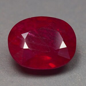 Könnte beinhalten: Ein tiefroter, oval geschliffener Edelstein mit facettierter Oberfläche. Der Stein zeigt einen lebendigen, gesättigten karmesinroten Farbton mit internen Einschlüssen und scharfen, geometrischen Lichtreflexen auf der polierten Oberfläche.