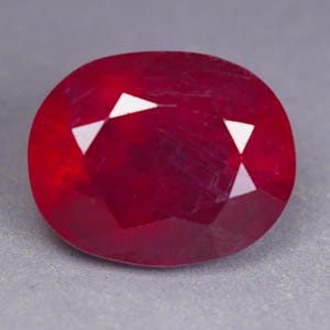May include: An oval-cut ruby gemstone with a deep, saturated red colour and a faceted surface. The stone displays internal clarity characteristics and sharp, geometric facets that reflect light, creating a brilliant appearance across the surface.