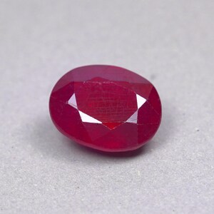 May include: A deep red, oval-cut gemstone with a faceted surface. The stone displays a rich, saturated crimson hue and exhibits multiple geometric facets that reflect light across its polished, translucent top and rounded edges.