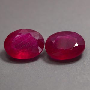 Puede incluir: Dos rubíes de corte ovalado con un color rojo intenso y saturado y superficies facetadas. Las piedras preciosas presentan un acabado pulido con inclusiones internas visibles y facetas que reflejan la luz en sus formas redondeadas.