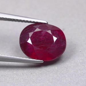 May include: A deep red, oval-cut gemstone with a faceted surface, held by a pair of silver-toned metal tweezers. The stone displays a rich, saturated crimson hue with visible internal inclusions and a polished, reflective finish throughout.